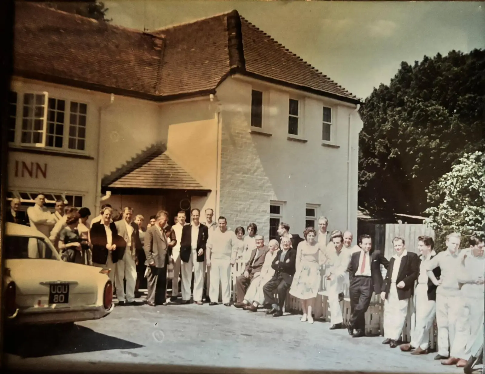 Colour photograph from around 1960 of a crowd of villagers, many in cricket whites, gathered outside the inn